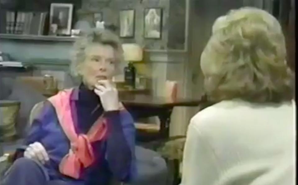 Katharine Hepburn and another person are in a room with bookshelves. Hepburn is seated, wearing a sweater and scarf, looking at the other person