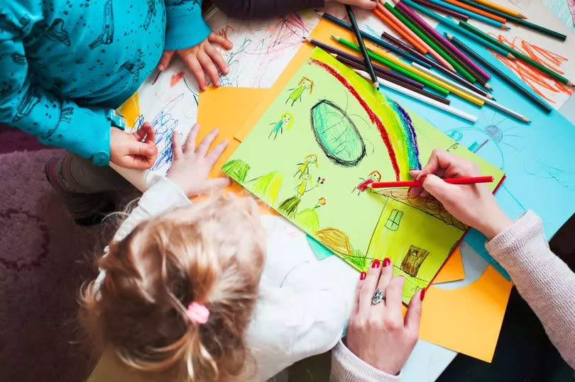 Mom with her little daughters and son together drawing a colorful picture of playing children using pencil crayons sitting at table. Photo from above