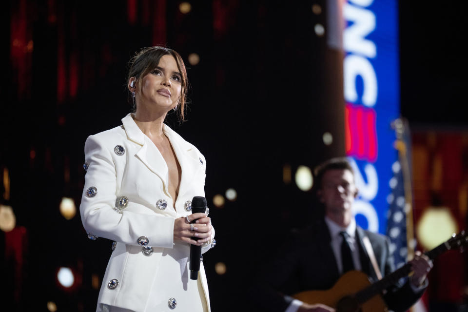 Maren Morris tritt in einem eleganten weißen Outfit mit Juwelenverzierungen auf der Bühne auf und hält ein Mikrofon in der Hand. Im Hintergrund spielt ein Gitarrist