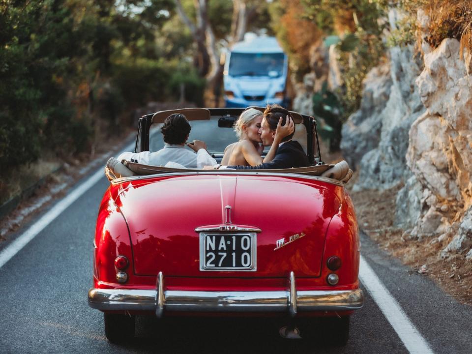 A couple kiss in a red convertible as they drive down a mountain.