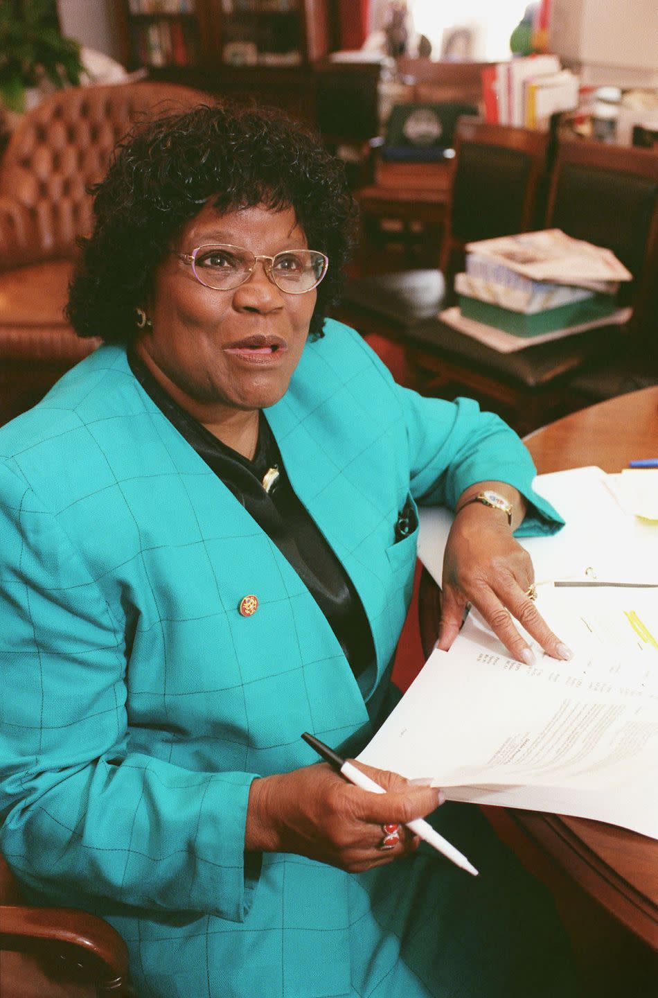 carrie meek looks to the right of the camera while sitting at a table with paperwork in front of her, she wears a teal blazer over a black blouse, glasses and jewelry