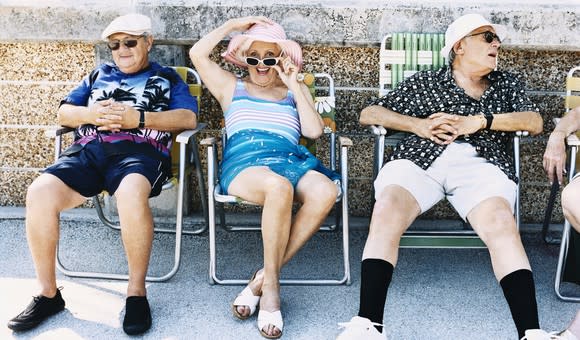 Three senior citizens in summer clothes sitting and chatting on folding lawn chairs.