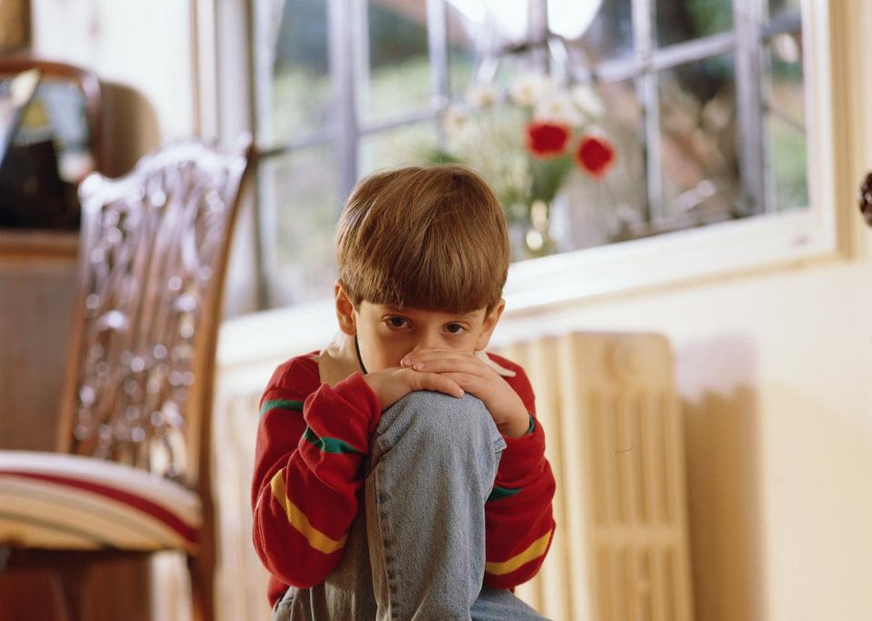 Child in a red top and jeans sitting on a step with knees up and resting head on hands, looking thoughtful