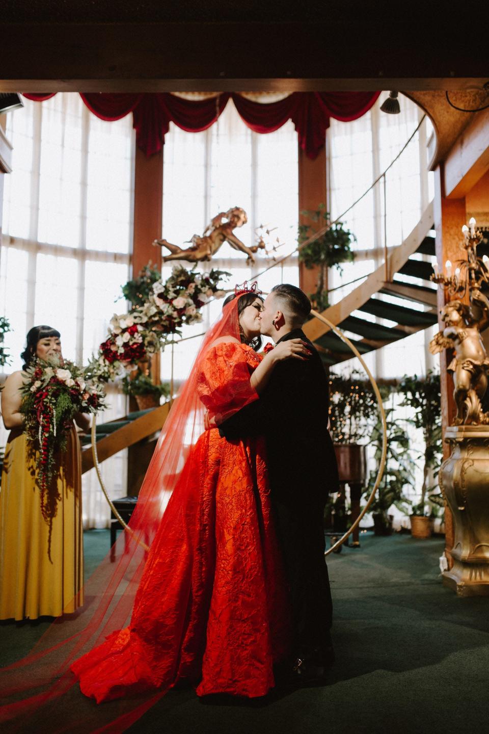 A woman in a red dress and a man in a black suit kiss during their wedding ceremony.