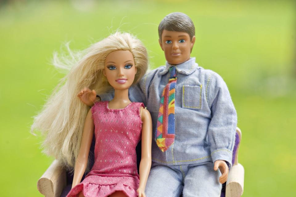 barbie and ken dolls sit on a couch, she wears a pink dress, he wears a denim shirt and tie with jeans