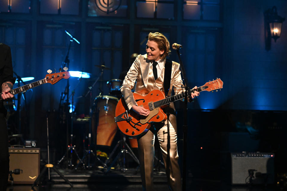 Person playing guitar on stage, dressed in a suit, with band members and instruments in background