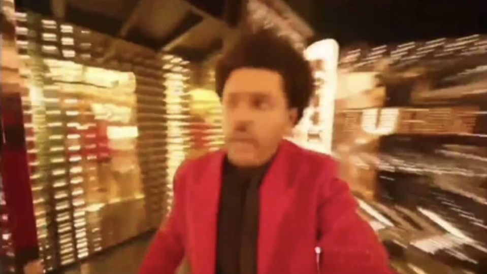 The Weeknd, wearing a stylish jacket, appears to be walking through a brightly lit, bustling modern setting. The image has a motion blur effect