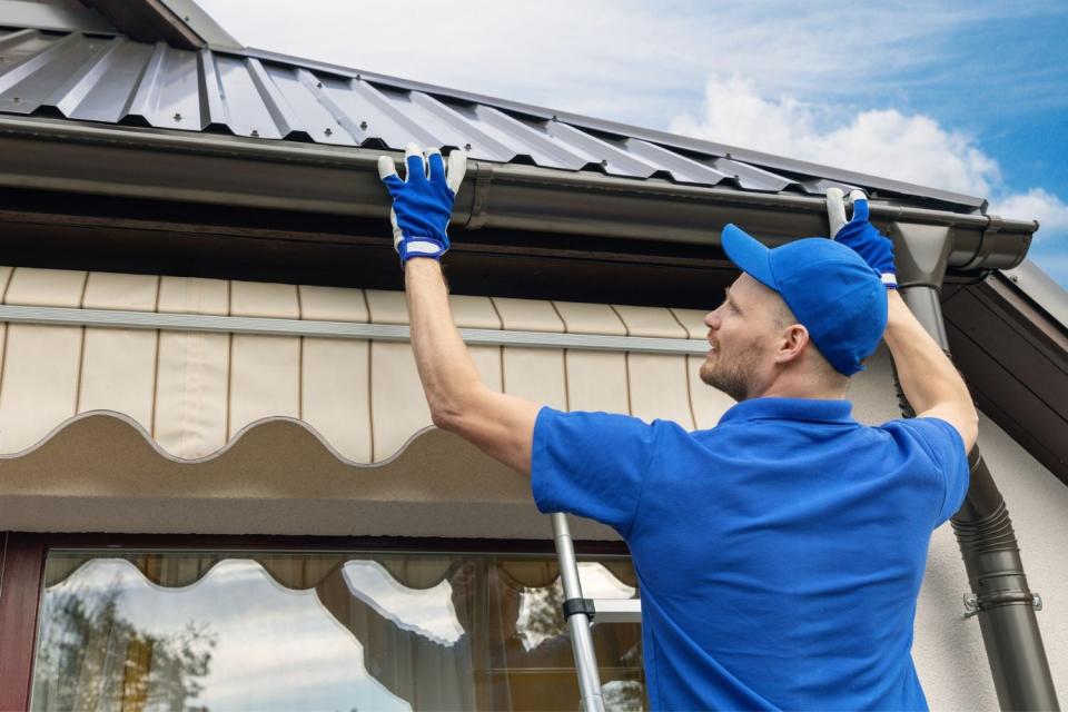 A man dressed in blue work clothes on a ladder installs a gutter guard.