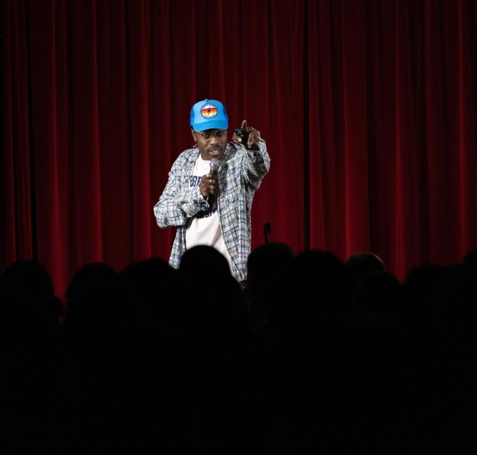 A man in a blue baseball cap onstage holds a microphone with one hand and gestures with the other