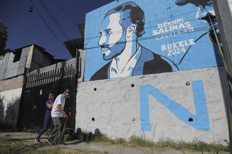 A woman guides an elderly man through the imperfections on a sidewalk past a New Ideas campaign mural promoting President Nayib Bukele who is running for re-election, in the Mejicanos suburb of San Salvador, El Salvador, Wednesday, Jan. 24, 2024. El Salvador will hold a general election on Feb. 4 where voters will elect a president, vice president and members of Congress. (AP Photo/Salvador Melendez)