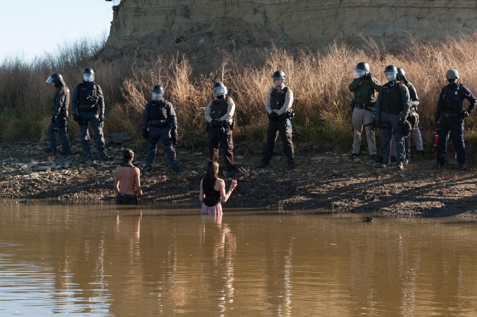 Two people stand in the water of a river while police officers guard the shore.