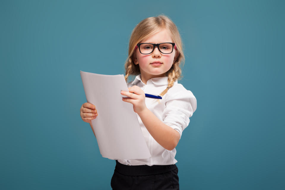 Young child wearing glasses holds papers and a pen, appearing focused and thoughtful against a simple backdrop