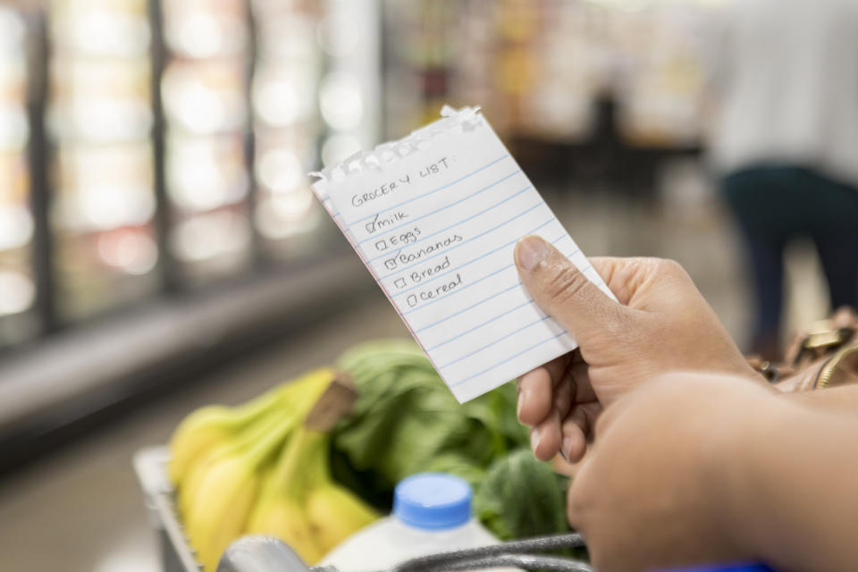 In this point of view photograph, an unrecognizable person, with only hands showing, holds a paper shopping checklist over a full shopping cart.  The customer is standing in the refrigerated foods aisle.
