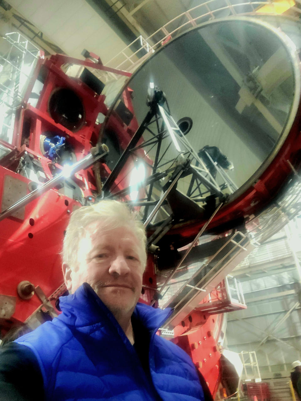 A man in a blue vest stands in a large room in front of a large piece of red equipment and a large, dark circular disc towering above it.