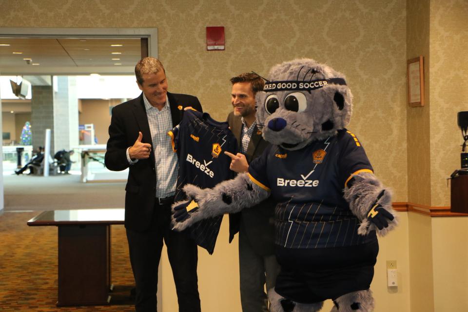 RIFC founder and chairman Brett Johnson, left, and Breeze Airways president Tom Doxey unveil the new professional soccer team's uniform with the help of team mascot Chip at an announcement Monday that Breeze will be the team's official airline partner.