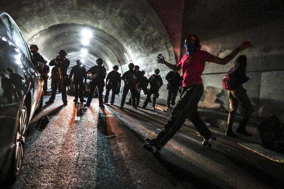 A woman lifts both arms as she roller skates in the street. Behind her are a row of police officers in riot gear.