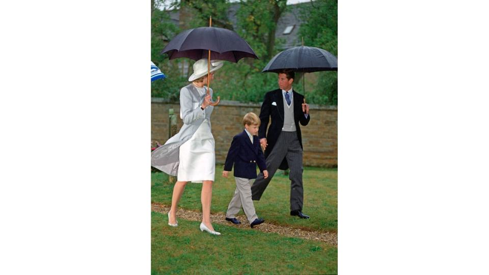 Princess Diana in a white dress and a grey coat under an umbrella with Charles and William