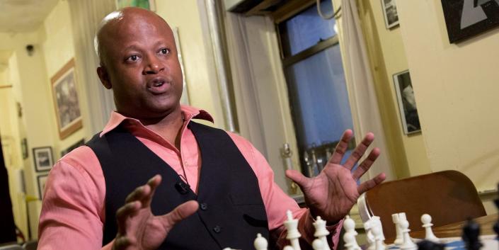 Maurice Ashley holds up his hands and speaks in front of a chess board during an interview.