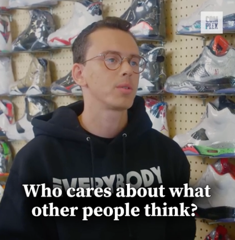 Logic stands in a sneaker store in a black hoodie with the text "EVERYBODY." Caption below reads, "Who cares about what other people think?"