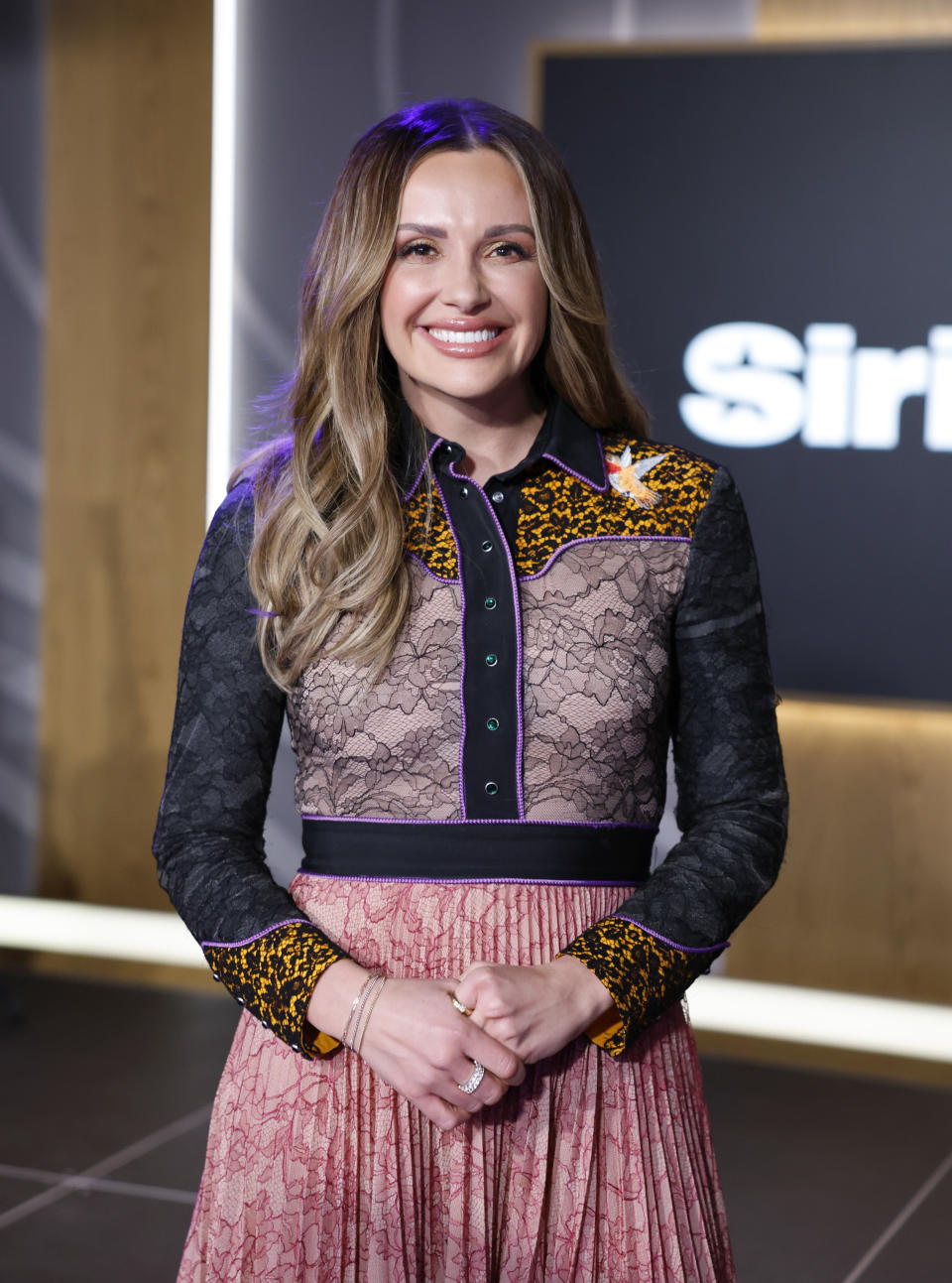 Carly Pearce smiles, wearing a lace blouse with leopard print details and a pleated skirt, at a SiriusXM event