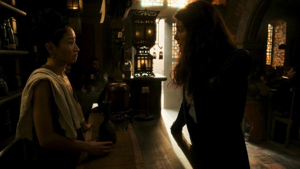 A shot of Min behind the bar speaking to Moiraine. Min has a white dish cloth over her shoulder and a shot of alcohol in her hands