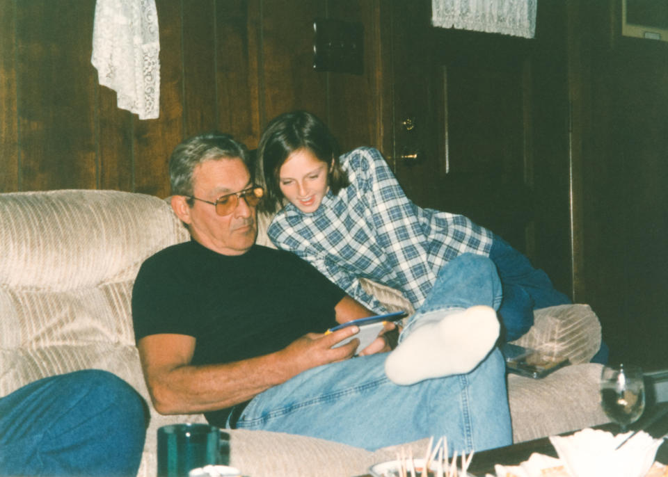 An older man and a young woman relax on a sofa. The man in sunglasses reads a document, while the woman in a plaid shirt leans over to look