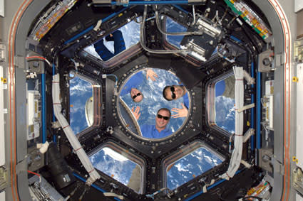 The three astronauts are outside the ISS wearing sunglasses. They are smiling and waving at the camera.