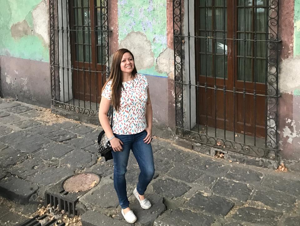 Woman wearing blue jeans, white shoes and a white speckled top standing on a stone street in front of a teal building with grates.