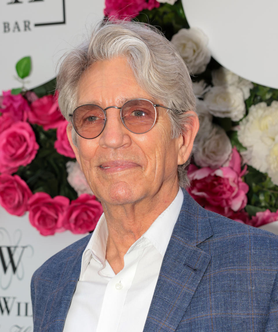 Eric Roberts at an event, wearing a blue blazer, white shirt, and round glasses, standing in front of a floral backdrop