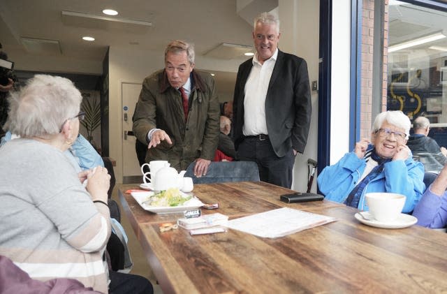 Nigel Farage gestures at a woman sitting at a table with some food and a tea pot as Lee Anderson watches on with his hands in his pocket and an elderly woman in a blue coat laughs 