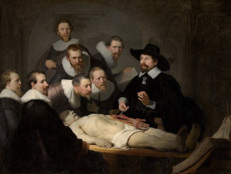 Six balding doctors gather round to watch a demonstration of a dissection.