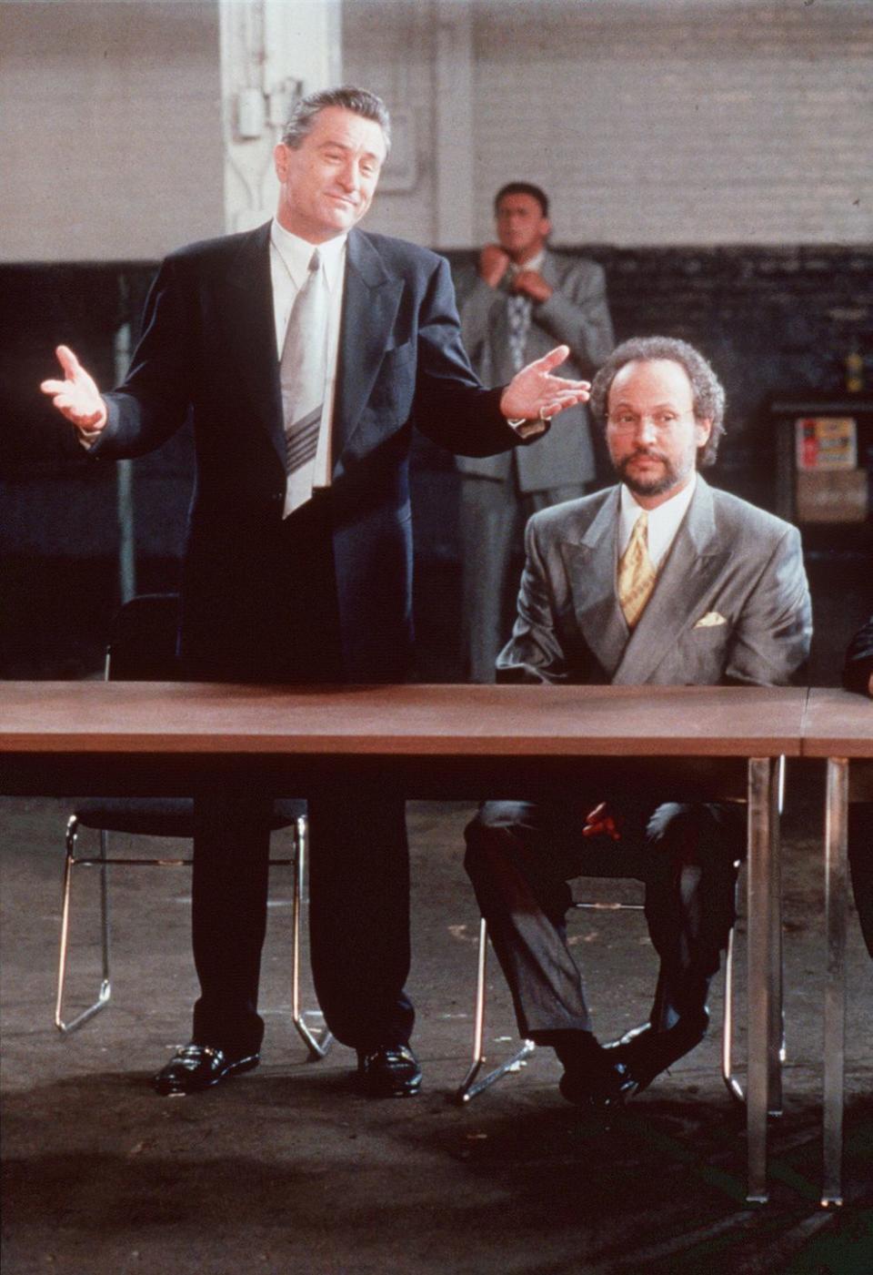 robert de niro wearing a black suit and tie and shrugging his shoulders, with billy crystal wearing a gray suit and tie and sitting next to him at a table