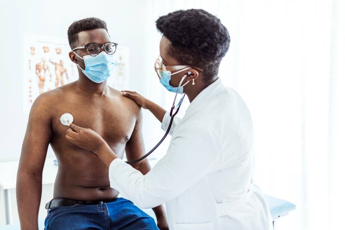 A doctor examines a male patient using a stethoscope in a medical office. Both are wearing masks. A poster with anatomical diagrams is visible in the background