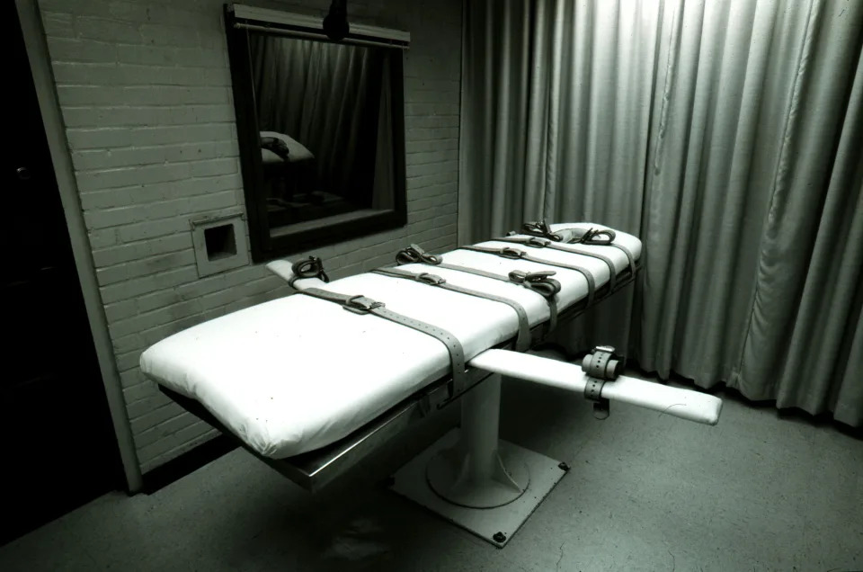 An execution bed sits empty next to a brick wall with a mirror and a floor to ceiling curtain.