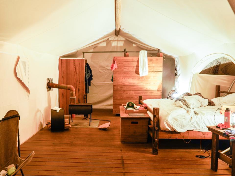 Inside a large tent with wood floors and white walls. On the left, theres a camping chair and a wood oven behind in. On the right, theres a king-sizeed bed, a chest, and night stands. The wood-walled bathroom is at the back of the tent.