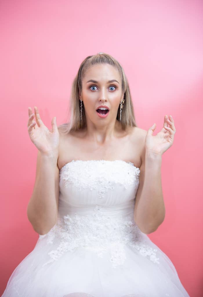 A woman with long blonde hair, wearing a strapless white wedding dress, looks surprised with her hands raised, posed against a plain background