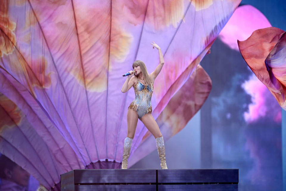 Taylor Swift performs on stage in a sparkling, sequined bodysuit and knee-high boots against a background of flowing, abstract floral patterns, atop a platform