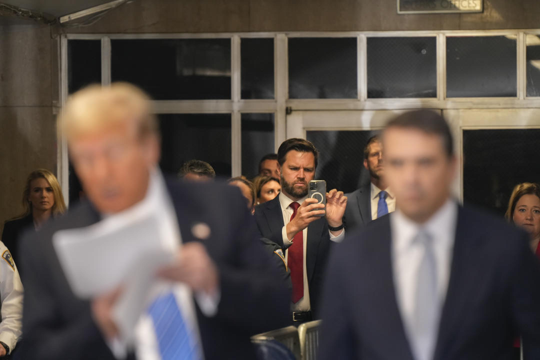 J.D. Vance, center, takes a photo with his cellphone as Donald Trump, blurred upfront, speaks to reporters at Manhattan criminal court.