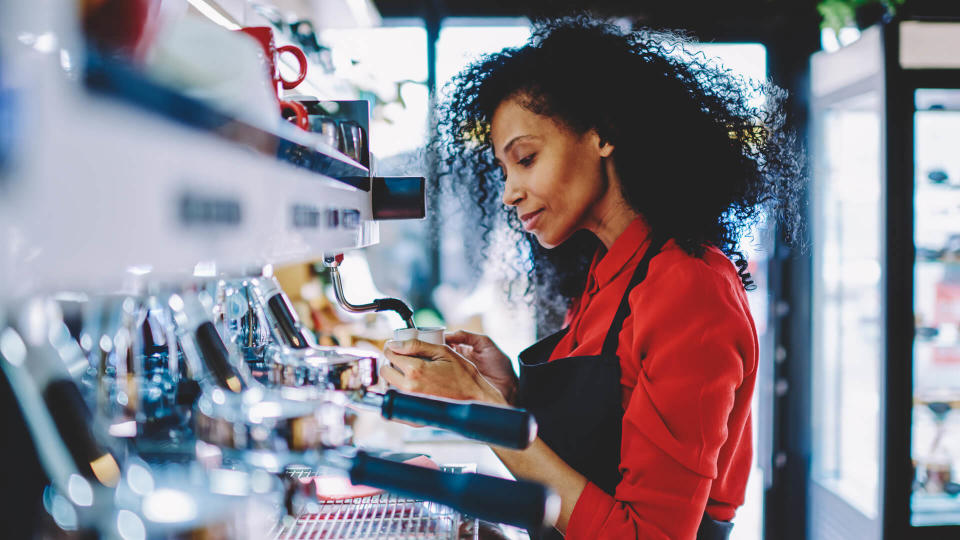 Professional female barista with dark skin standing at bar on working place and preparing tasty beverage on order.