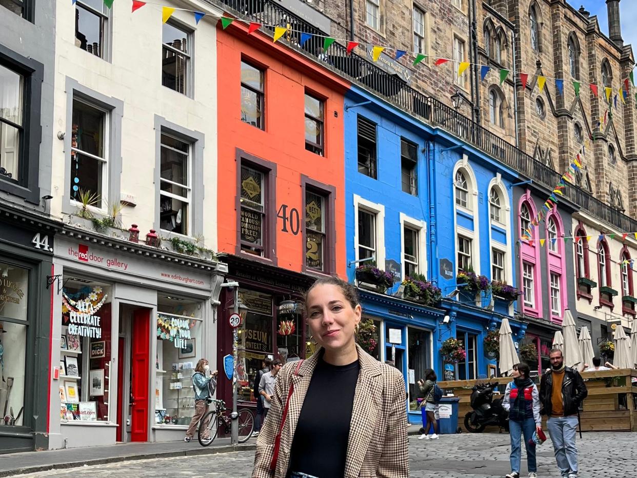 Dasha stands in front of a row of colorful buildings in Edinburgh. Behind her, people are walking around near the shops.