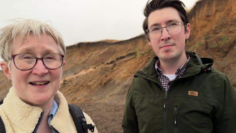 Sarah Chase and Will Chase standing on the beach with the sand-coloured cliffs in the background. Sarah has a cream fleece jacket, short grey hair and glasses. Jamie has is wearing a green jacket and has short dark hair and glasses