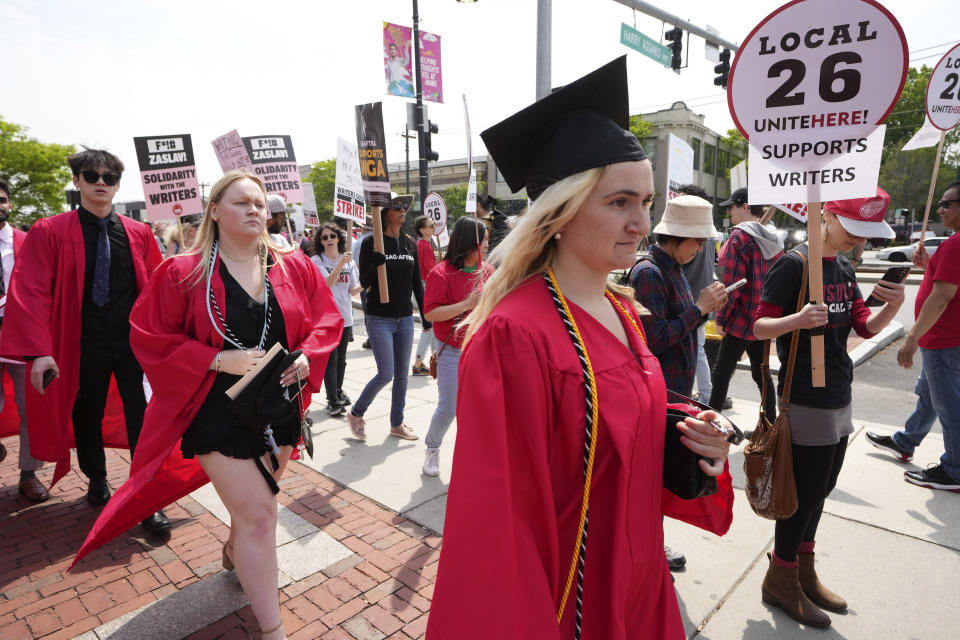 People dressed in commencement gowns, front, walk past protesters supporting the Hollywood writers' strike outside an entrance to Boston University commencement ceremonies, Sunday, May 21, 2023, in Boston. David Zaslav, president and CEO of Warner Bros. Discovery, delivered an address during the ceremonies Sunday. (AP Photo/Steven Senne)