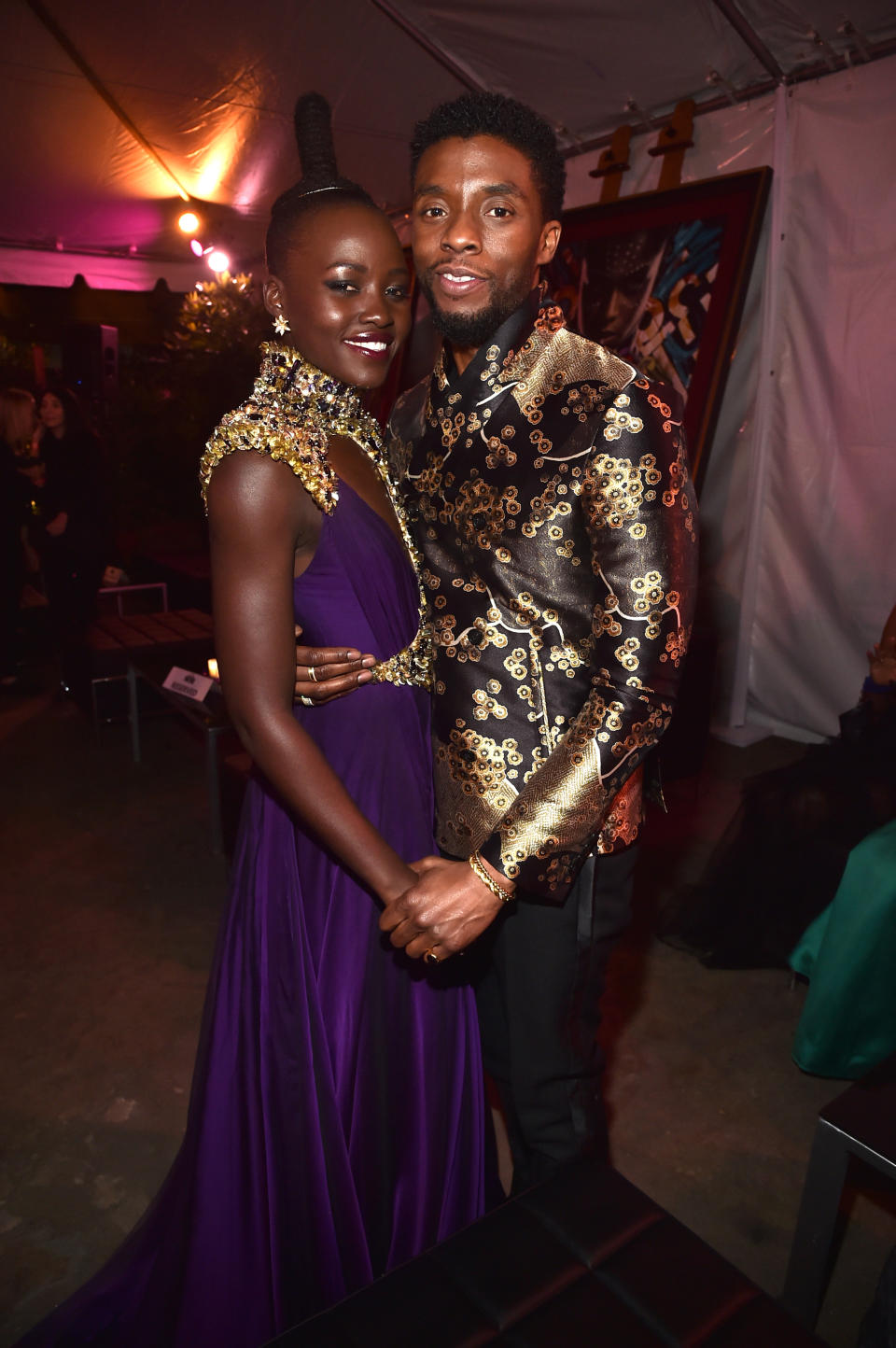 Lupita Nyong'o in a flowing gown with gold accents poses with Chadwick Boseman, who is wearing an ornate patterned jacket, on a red carpet event