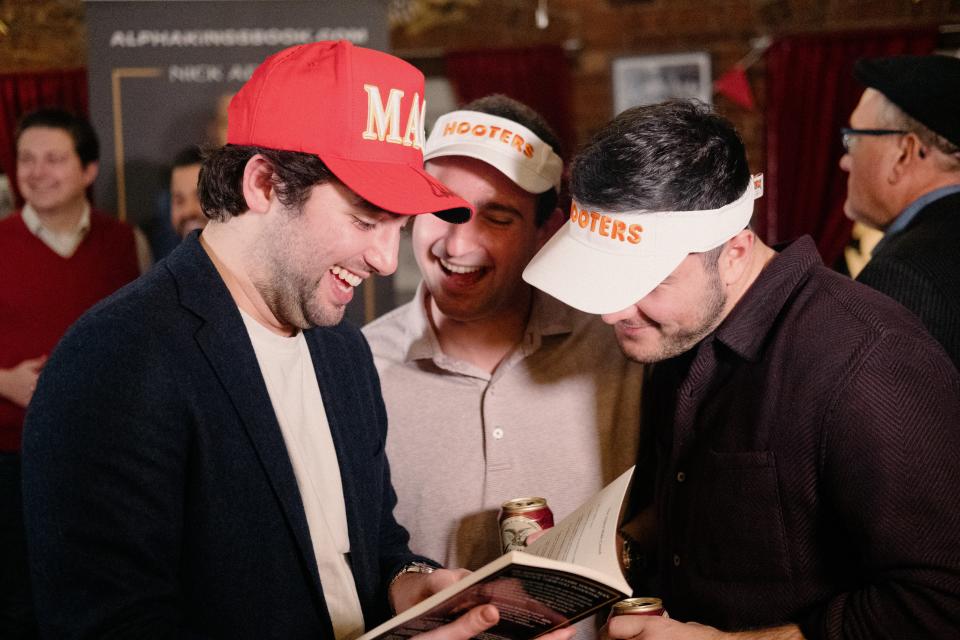 A trio of men, who are laughing and wearing Hooters and MAGA hats, thumb through Nick Adams' new book at the Young Republican event.