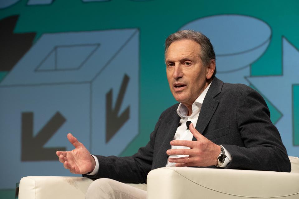 Howard Schultz sitting in a chair and gesturing with his hands while speaking to a crowd.