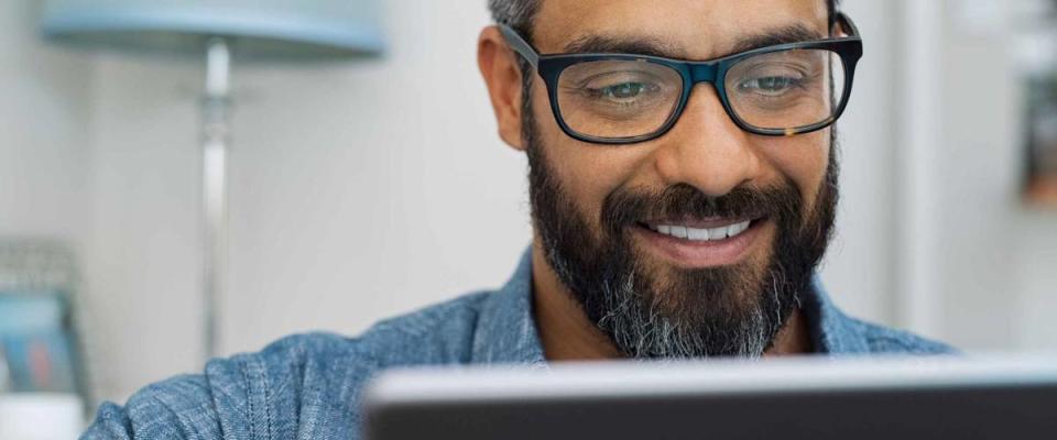 Relaxed mature man at home using digital tablet. Handsome hispanic man using laptop on sofa. Confident multiethnic guy with spectacles and beard using digital laptop.