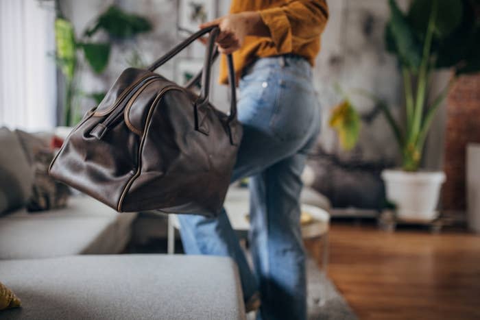A person in casual jeans and a top is picking up a large brown leather bag from a couch in a living room filled with plants