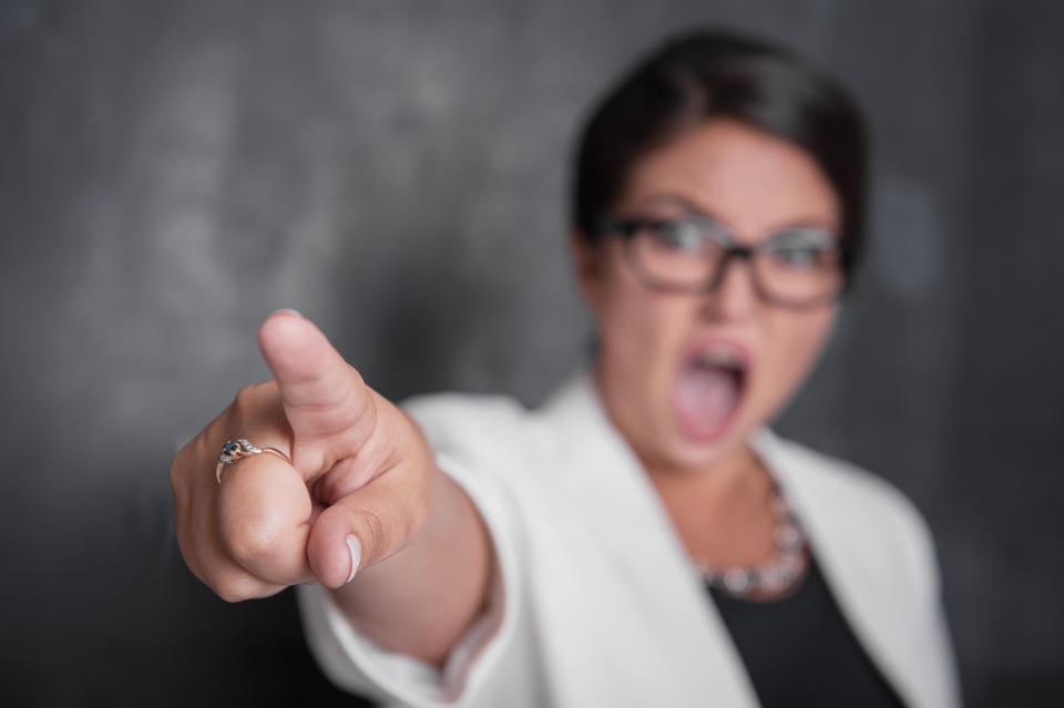 A person with glasses, wearing a blazer and a necklace, points aggressively toward the camera, mouth open as if yelling