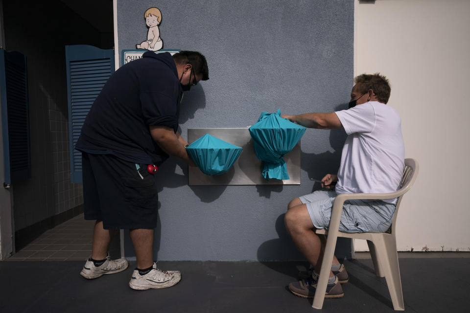 Adventure City amusement park employees, Michael Santy, left, and Clay Stark cover two water fountains with plastic sheets to prevent people from drinking from them as part of coronavirus safety measures ahead of the park's reopening in Anaheim, Calif., Wednesday, April 14, 2021. The family-run amusement park that had been shut since March last year because of the coronavirus pandemic reopened on April 16. (AP Photo/Jae C. Hong)