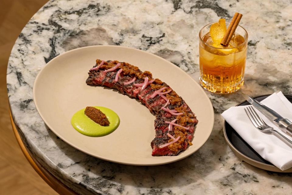 A plate of thin-sliced skirt steak with green tahini sauce, sitting on a marble tabletop next to an orange cocktail.